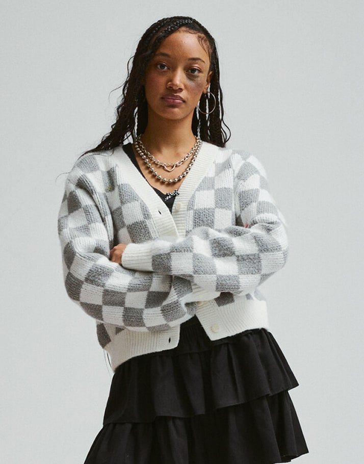 A young woman poses with her arms crossed wearing the Evelyn Cardigan Sweater in a photography studio.