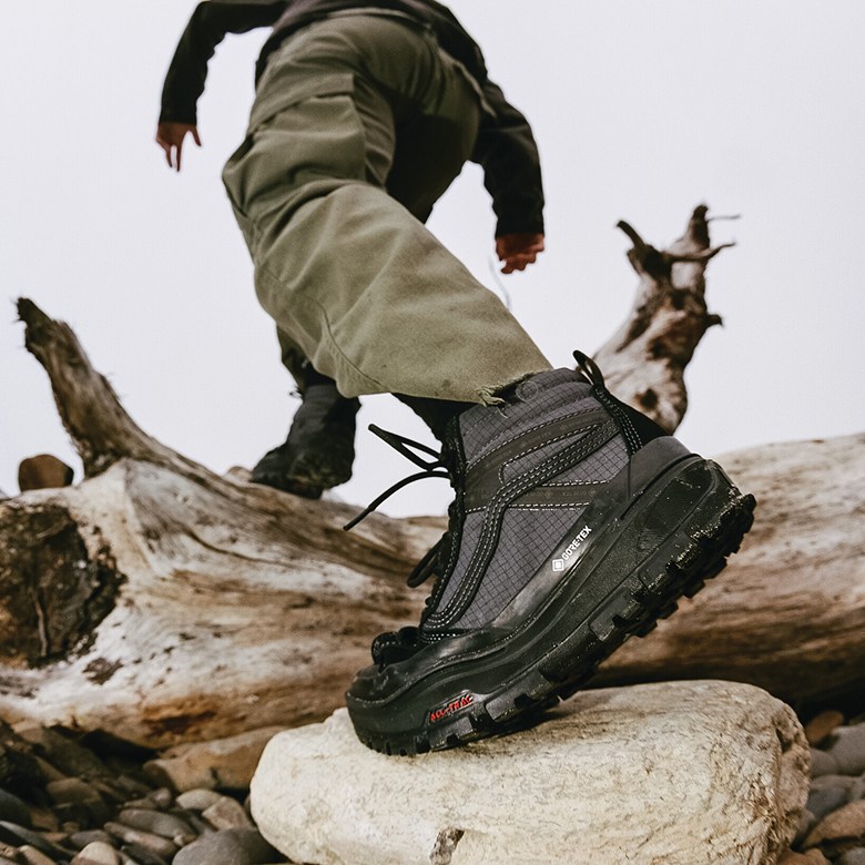 Un gros plan de quelqu'un portant le noir Sk8-Hi GORE-TEX marchant sur des rondins et des rochers à l'extérieur.