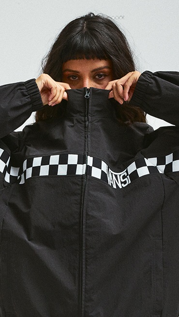 A young woman pulls the collar of the black Ronnie Track Jacket over her face in a photography studio.