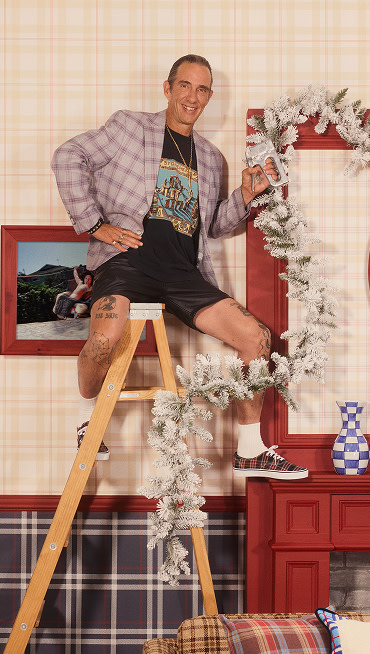Artist Cali DeWitt sits on a ladder stapling tinsel and wearing plaid Authentic on a sitcom set.