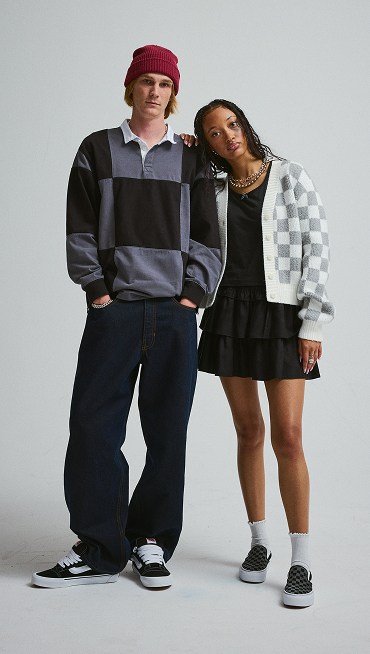 A young man in a gray and black Grant Checkerboard Rugby shirt stands next to a young woman in a Evelyn Cardigan Sweater in a photography studio.
