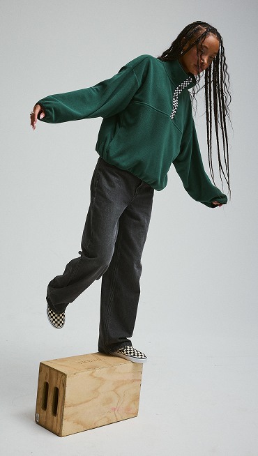 A young woman stands on a wooden box wearing the Emerald Polar Sweatshirt and Checkerboard Classic Slip-On in a photography studio.