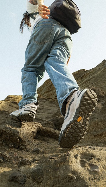 A man walking up a rocky face with white and blue MTE Crosspath shoes and blue jeans.