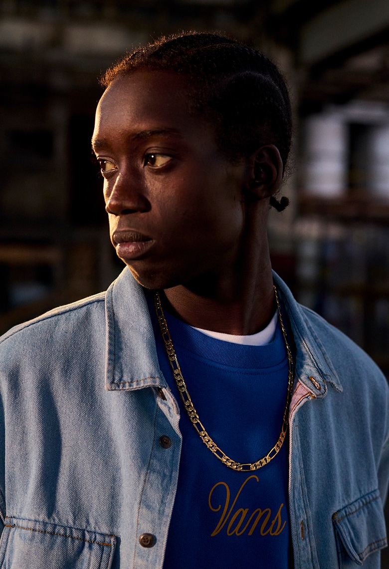 A man with posing and wearing a gold chain over a blue shirt that says Vans in cursive under a denim overshirt.