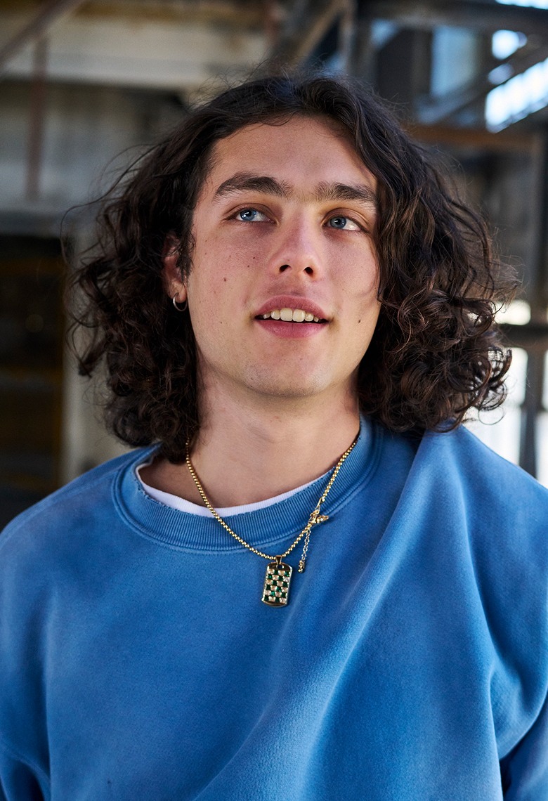 A man with long brown hair and blue eyes wearing a blue crewneck sweatshirt and a gold chain.