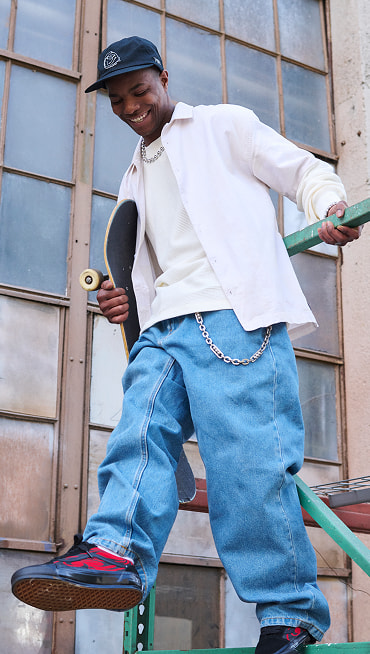 A man holding a skateboard on metal scaffolding wearing red and black flame prints on the Premium Old Skool Music Collection.