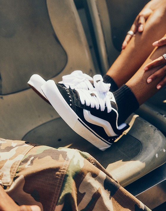 Close-up of a person wearing Rhinestone Black Knu Skool shoes, resting their feet on stadium-style seats.