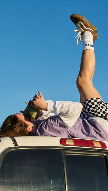 Person lying on a vehicle roof wearing checkered shorts and Old Skool shoes in Sepia Brown, holding a camera. One leg is raised against a clear blue sky.