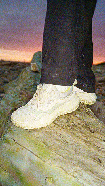 Person wearing MTE UltraRange shoes in white, and dark pants, stands on a large rock outdoors at sunset.