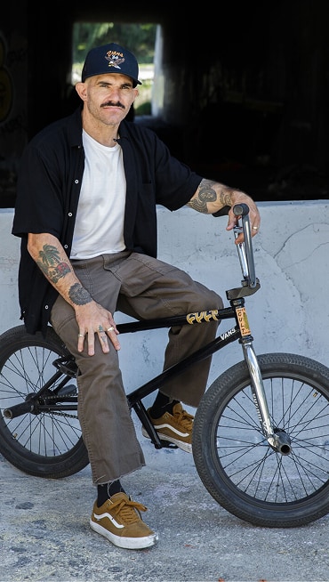 A man with tattoos and a mustache is sitting on a BMX bike while wearing Golden Brown BMX Old Skool shoes in front of a tunnel.
