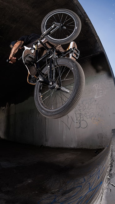 BMX rider performs an aerial trick against a graffiti-covered ramp under an overpass.