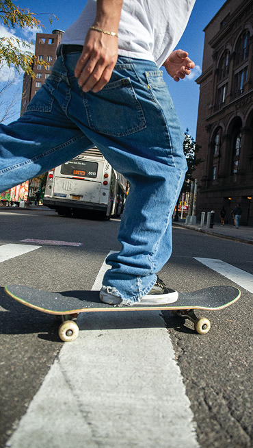 A skater skates across a street, wearing a white tee, baggy jeans, and Skate Old Skool 36+ sneakers in Dan Green.