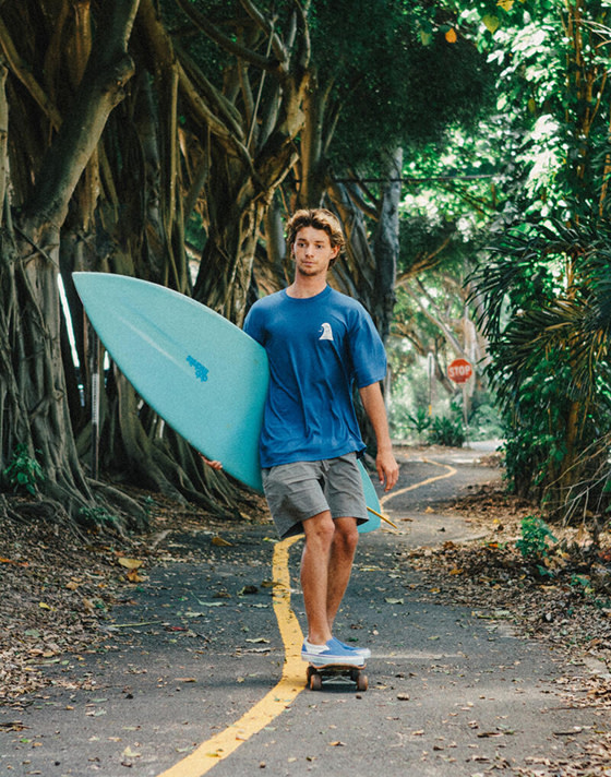 Surfer Tosh Tudor skates down a concrete trail with large trees and foliage all around. Tudor carries a blue surfboard and wears a Thalia Surf x Vans t-shirt in blue, shorts, and Slip-On Reissue sneakers in Thalia Blue.