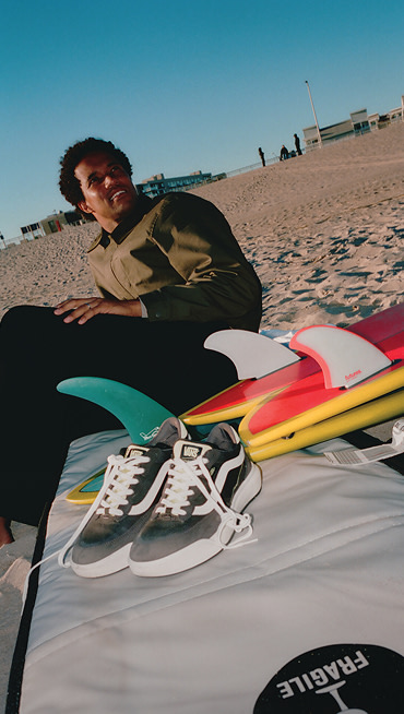 A man relaxes on a beach next to his surfboard and gray UltraRange 2.0 shoes.