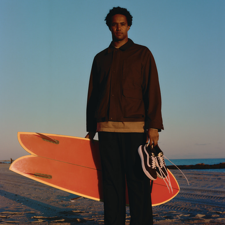 A man poses on a beach at golden hour holding his surfboard and Black and White UltraRange2.0.