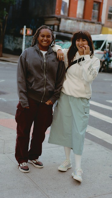 Two women pose on a street corner. One wears a Vans hoodie and red cargo pants while the other is in a white Vans hoodie and a skirt.