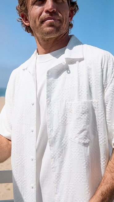 A close-up of a man's white Wayside Eyelet Woven Shirt over a t-shirt with the beach behind him.
