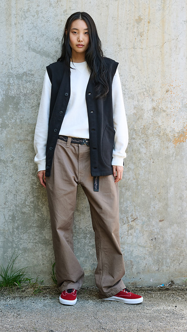 A woman poses against a concrete wall in a black vest, baggy brown pants, and red Curren shoes.