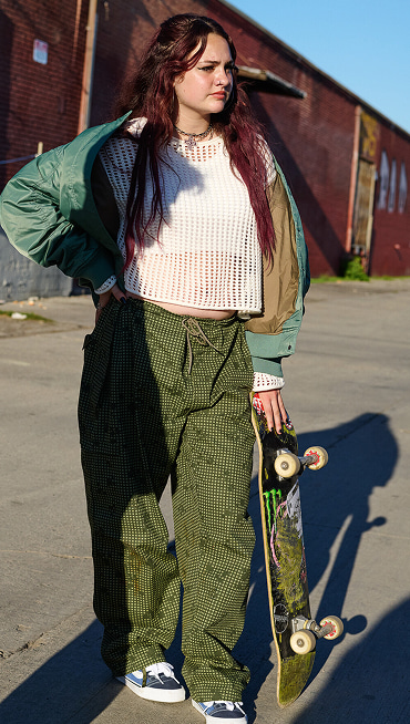 A woman looks into the distance and holds a skateboard wearing a white mesh top and baggy green pants in a parking lot.