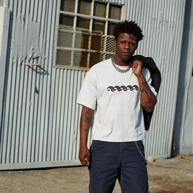 A man in a white Vans t-shirt with a silver chain and jacket over his shoulder looks at the camera with a rusty building behind him.